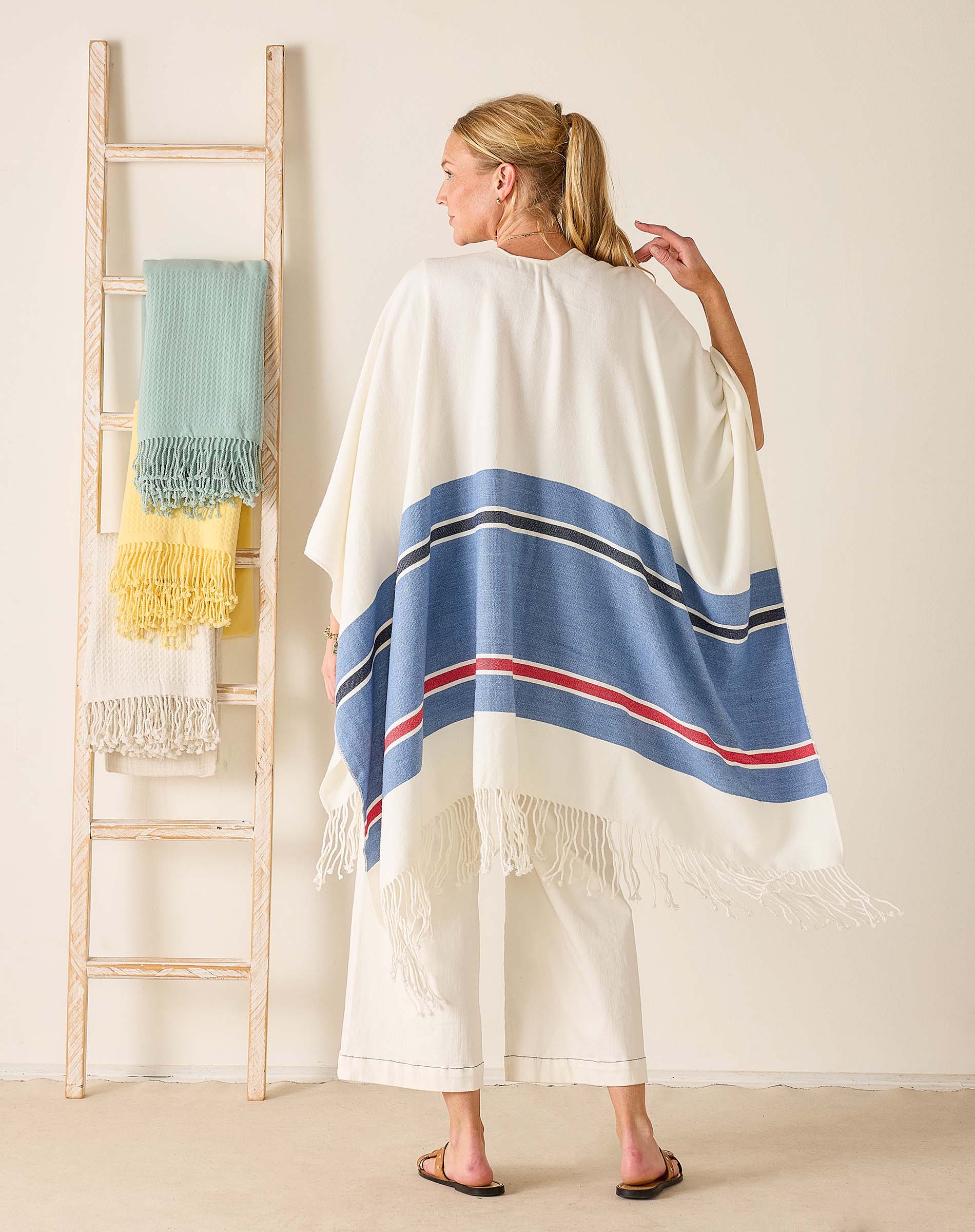 Woman wearing a white poncho with blue and red stripes, standing in front of a ladder with colorful cloths.