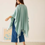 Woman wearing a light green poncho with fringe details, standing against a plain background.