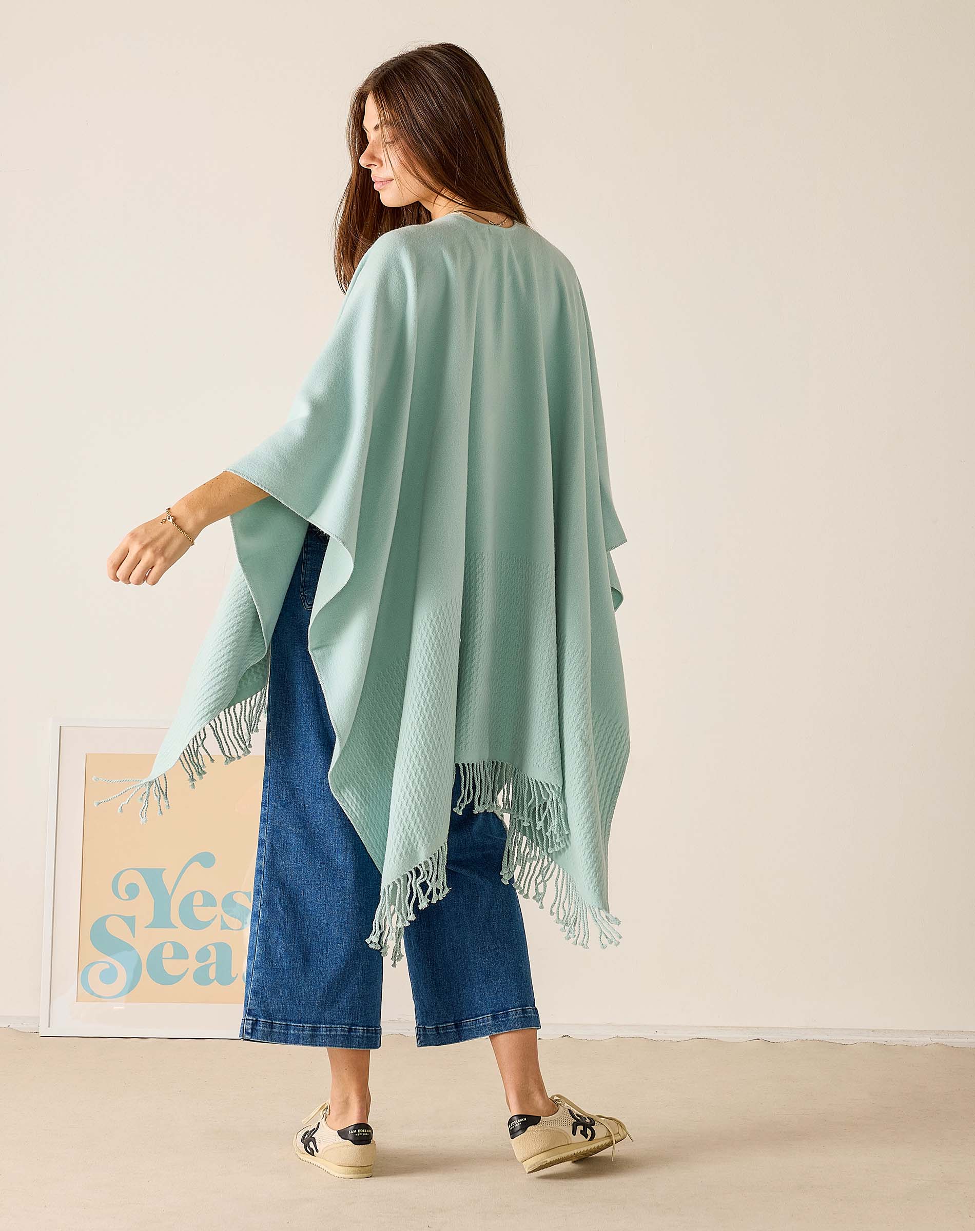 Woman wearing a light green poncho with fringe details, standing against a plain background.