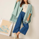 Woman wearing a light blue shawl and blue jeans against a white wall with a 'Yes Sea' poster.