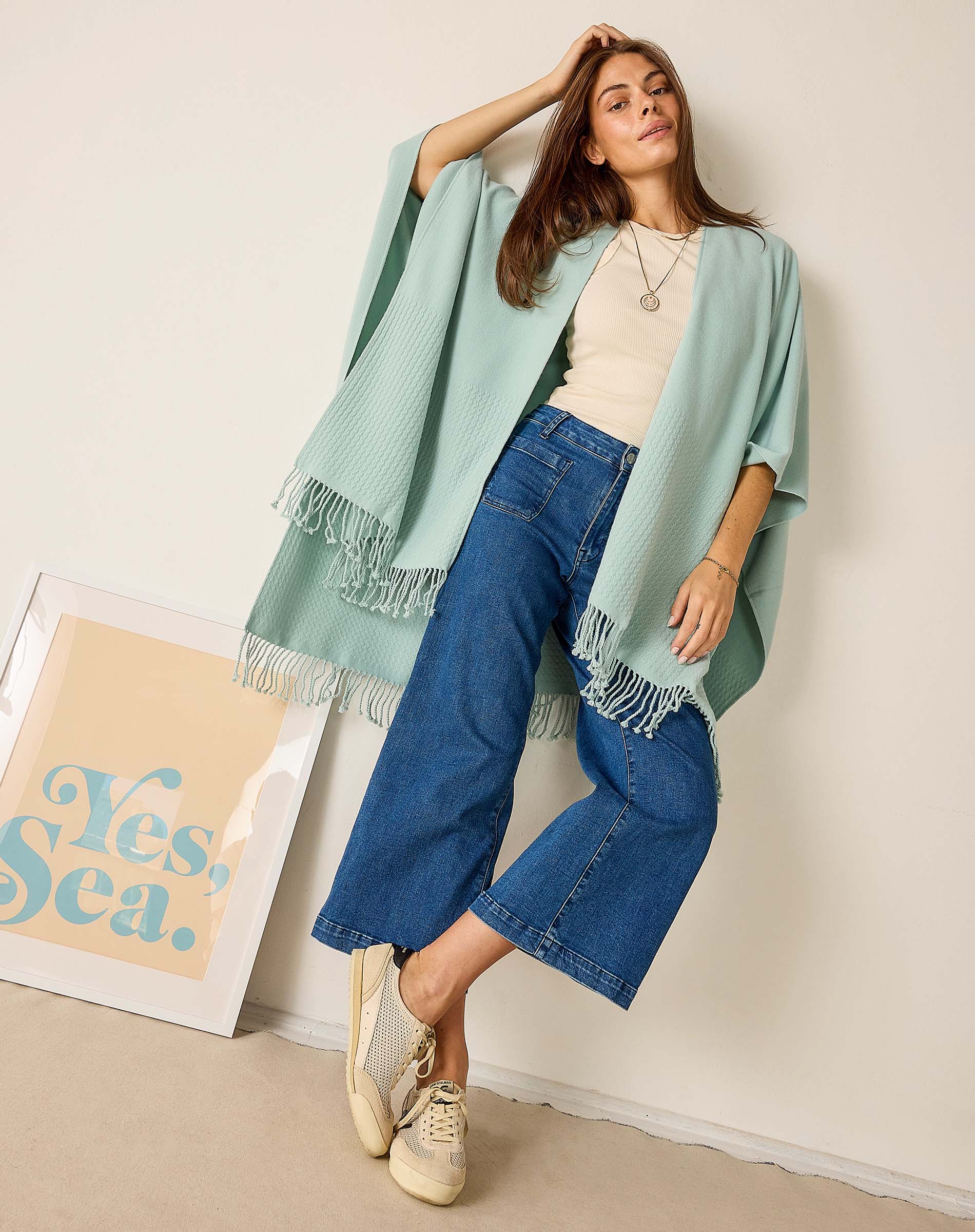 Woman wearing a light blue shawl and blue jeans against a white wall with a 'Yes Sea' poster.
