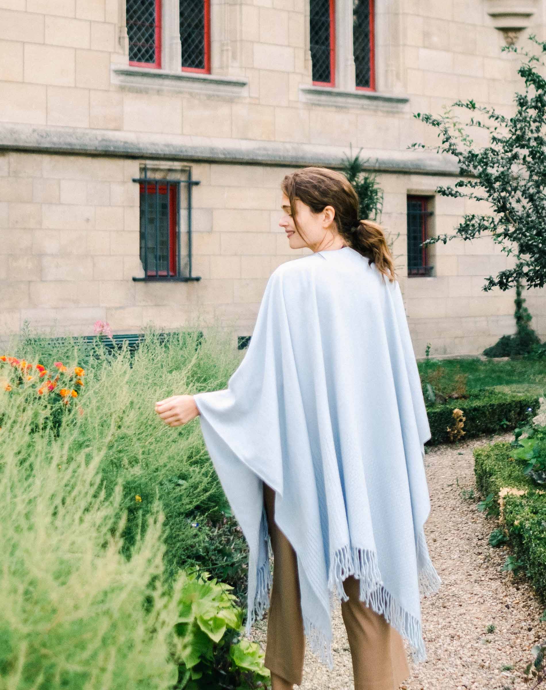 Woman wearing a light blue poncho in a garden setting with a building in the background