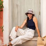 Woman sitting on a step wearing a navy top, white pants, and a hat with a straw bag.