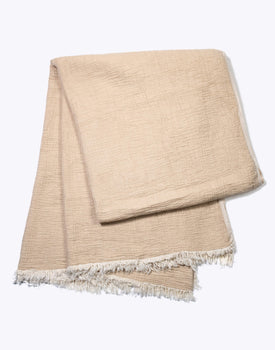 light beige gauze blanket with white fringe detailing laying folded on a white background
