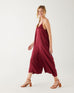walking woman wearing mersea coco satin jumpsuit in garnet against white backdrop