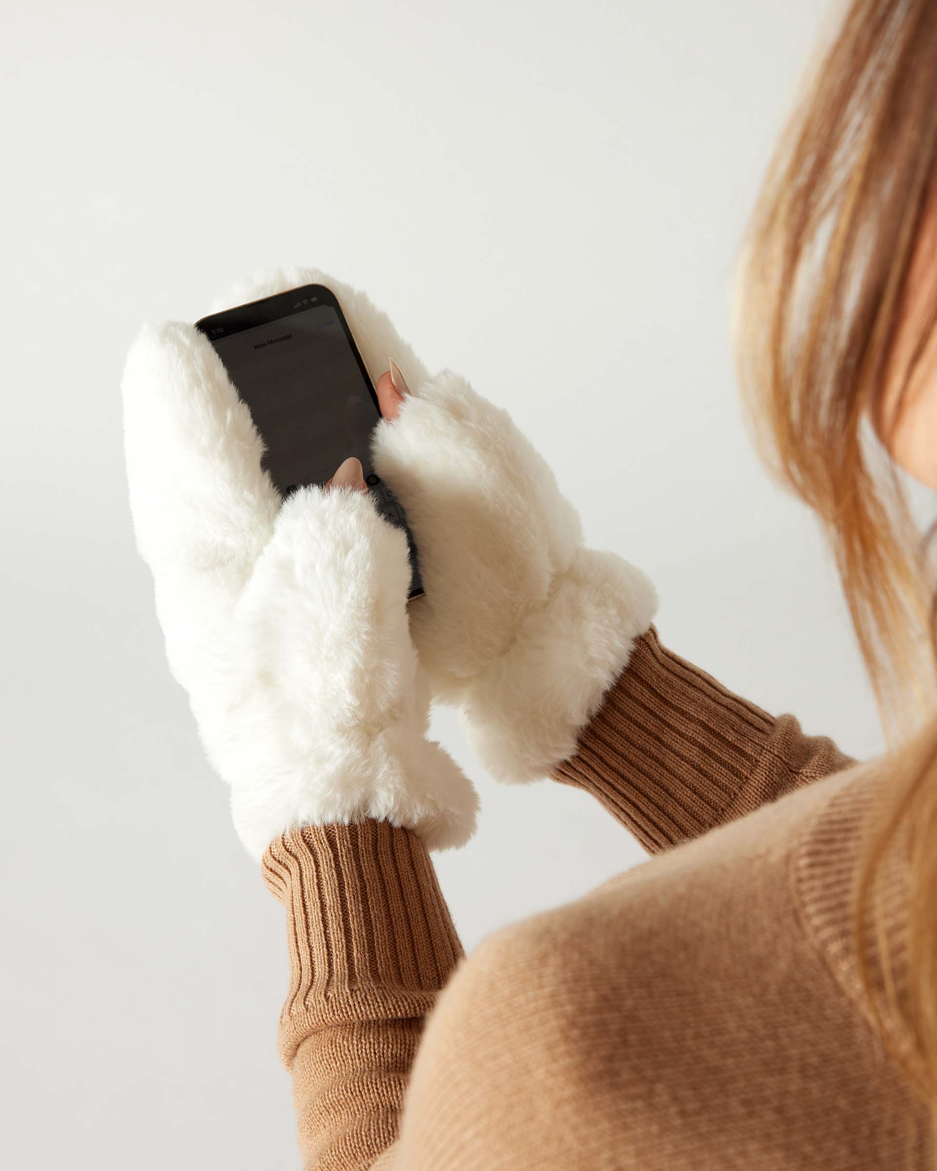 closeup of woman wearing mersea cozy cabin faux fur mittens in whisper white with exposed thumbs using phone