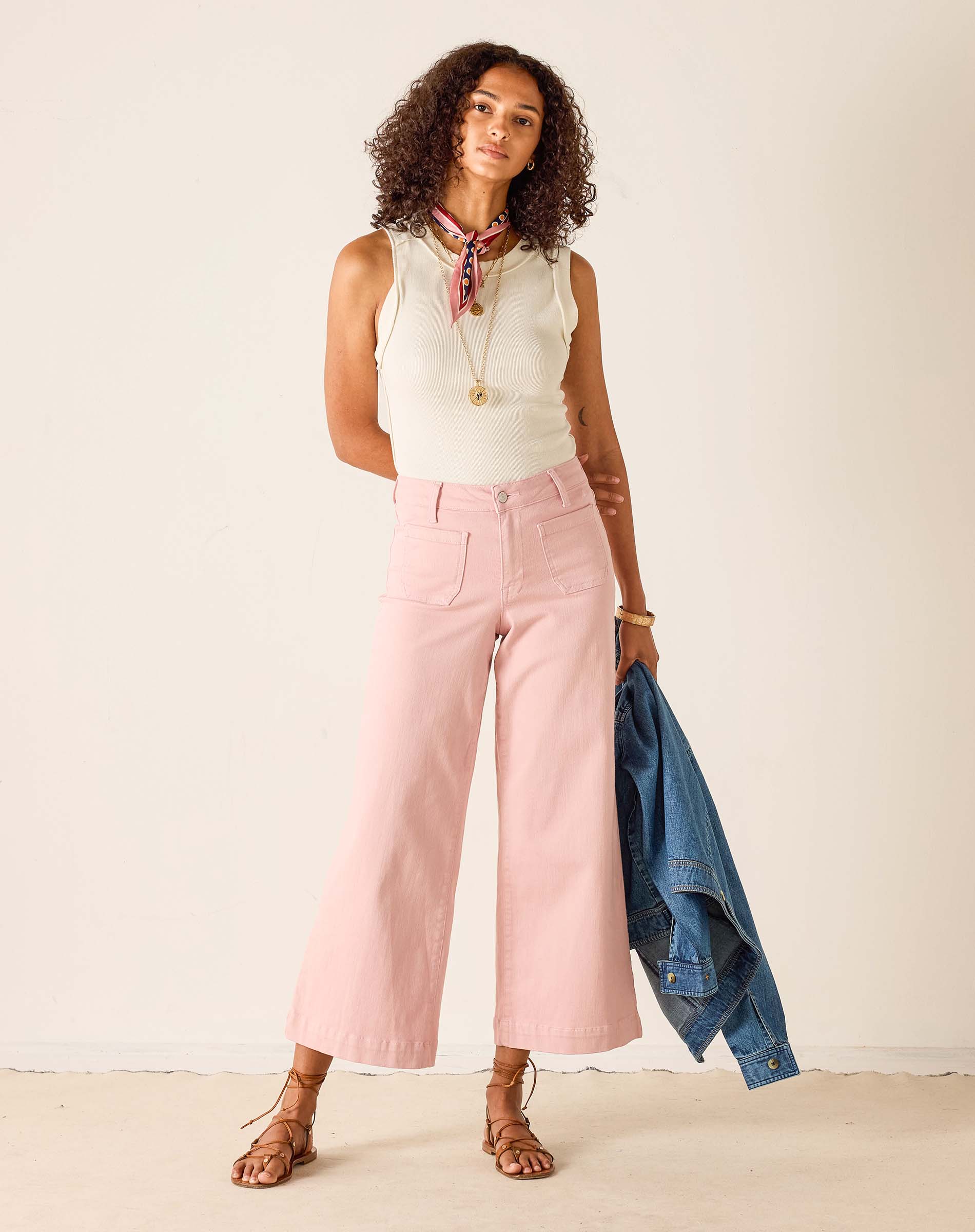 Woman wearing a sleeveless top, pink pants, and holding a denim jacket against a plain background