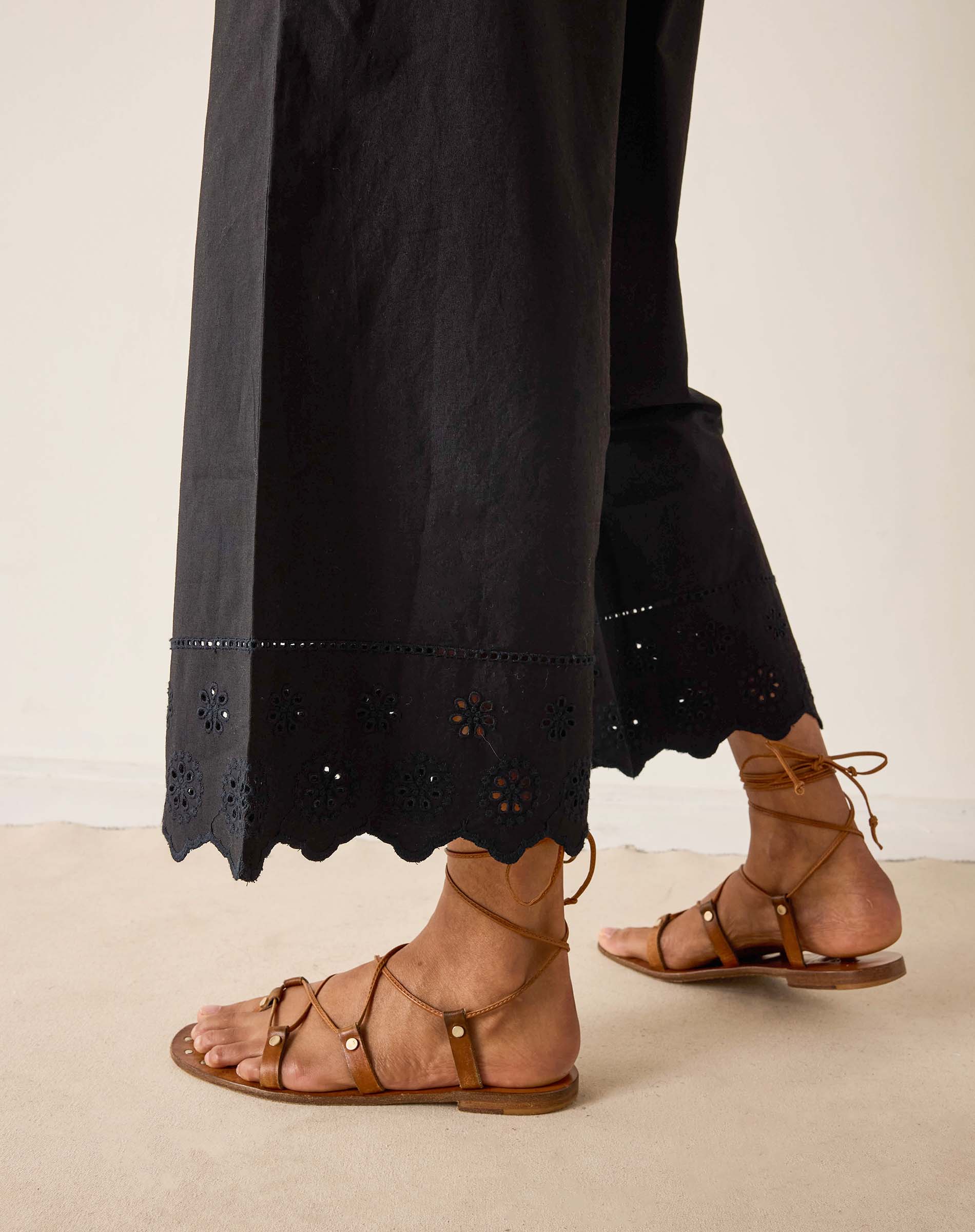 Person wearing black pants with eyelet details and brown sandals on a neutral background