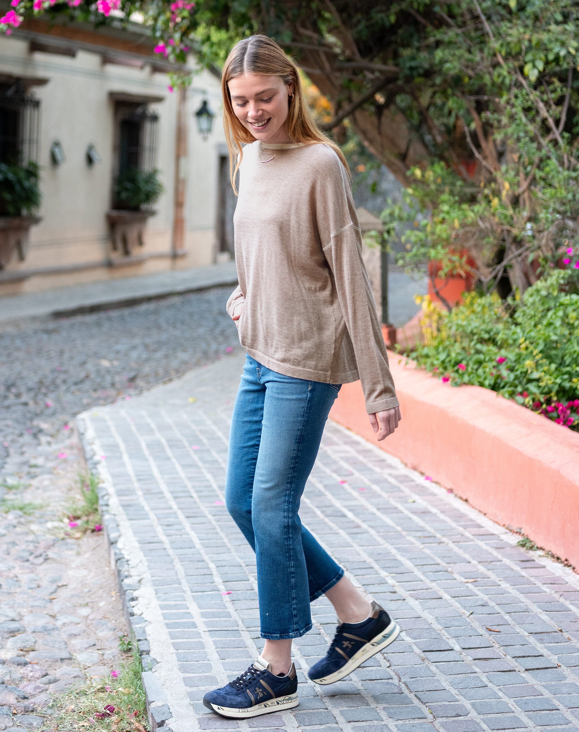 Woman walking on a cobblestone street wearing a beige sweater and blue jeans.