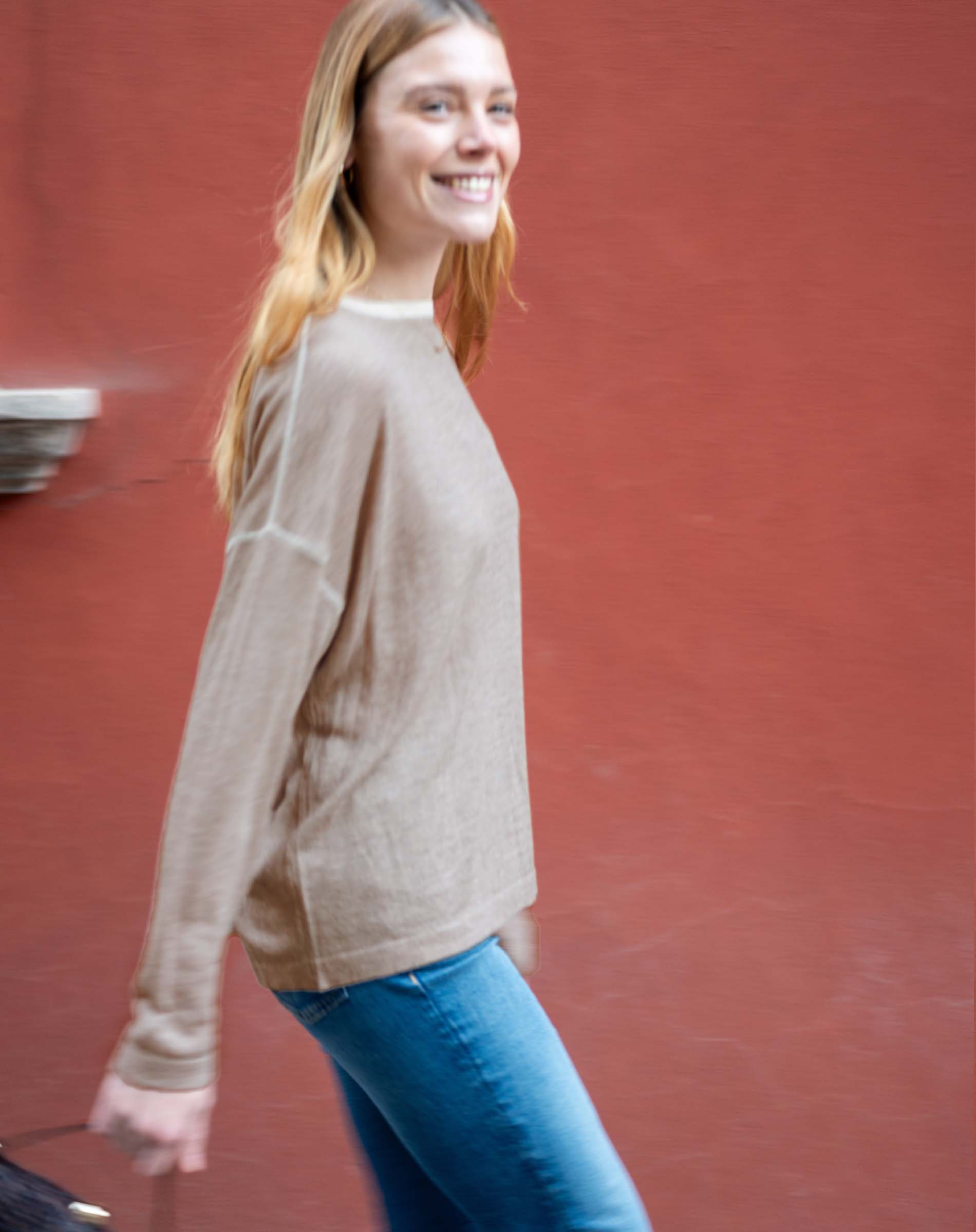Woman wearing a beige sweater and blue jeans against a red wall