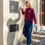 Woman in a burgundy sweater and blue jeans standing against a beige wall with decorative grates.