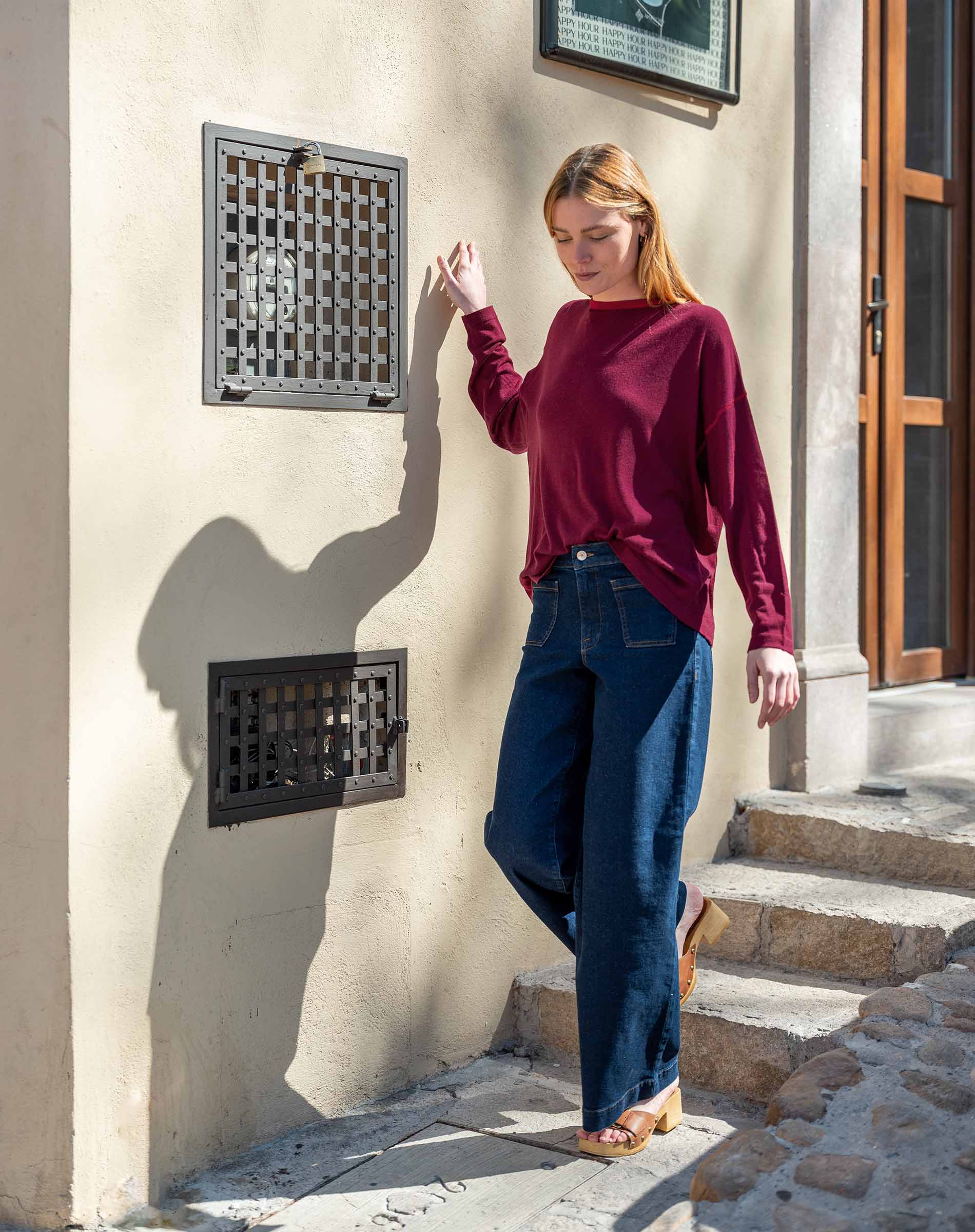 Woman in a burgundy sweater and blue jeans standing against a beige wall with decorative grates.