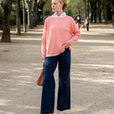 Woman wearing a pink sweater and blue jeans standing on a street with trees in the background