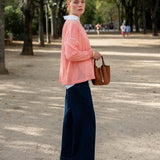 Woman in a pink jacket and blue pants standing on a tree-lined street.
