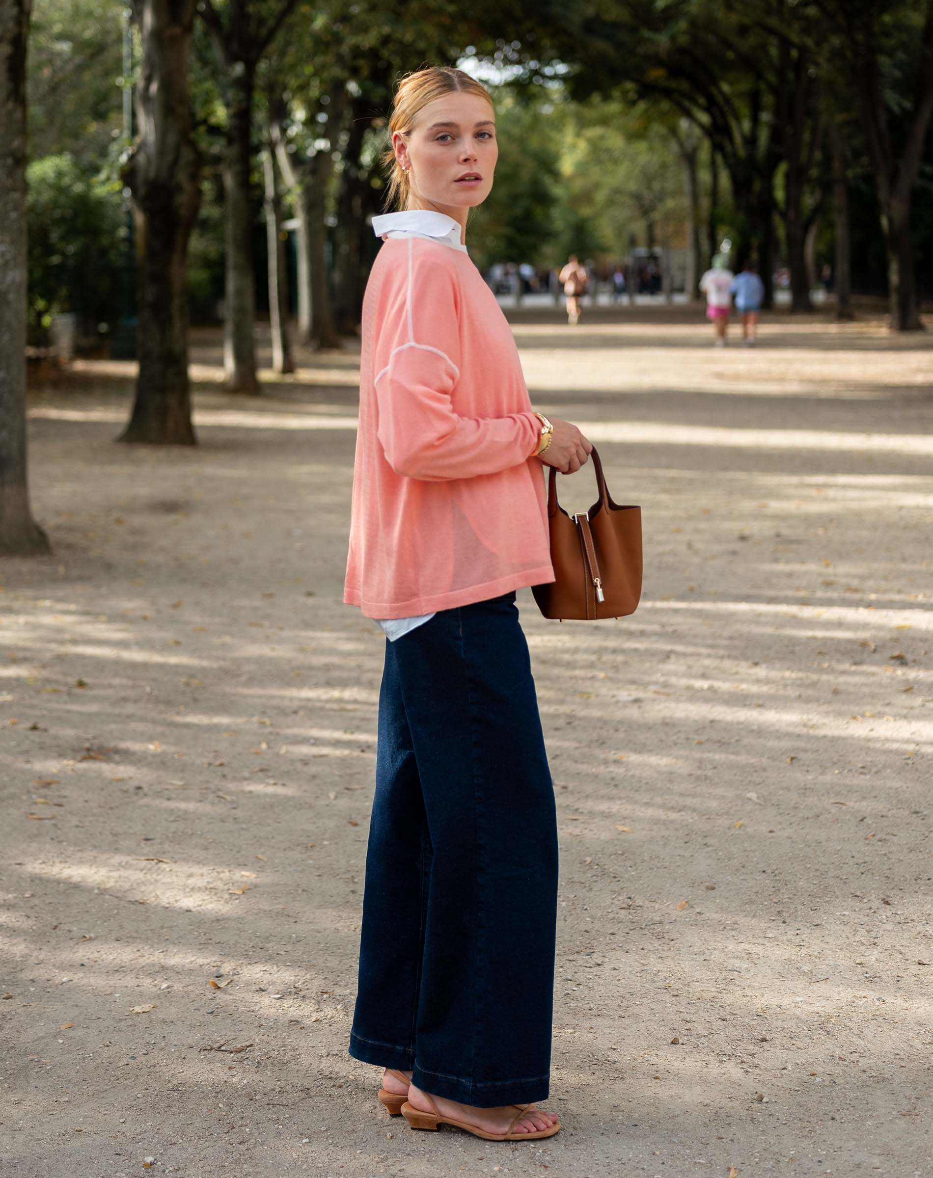 Woman in a pink jacket and blue pants standing on a tree-lined street.