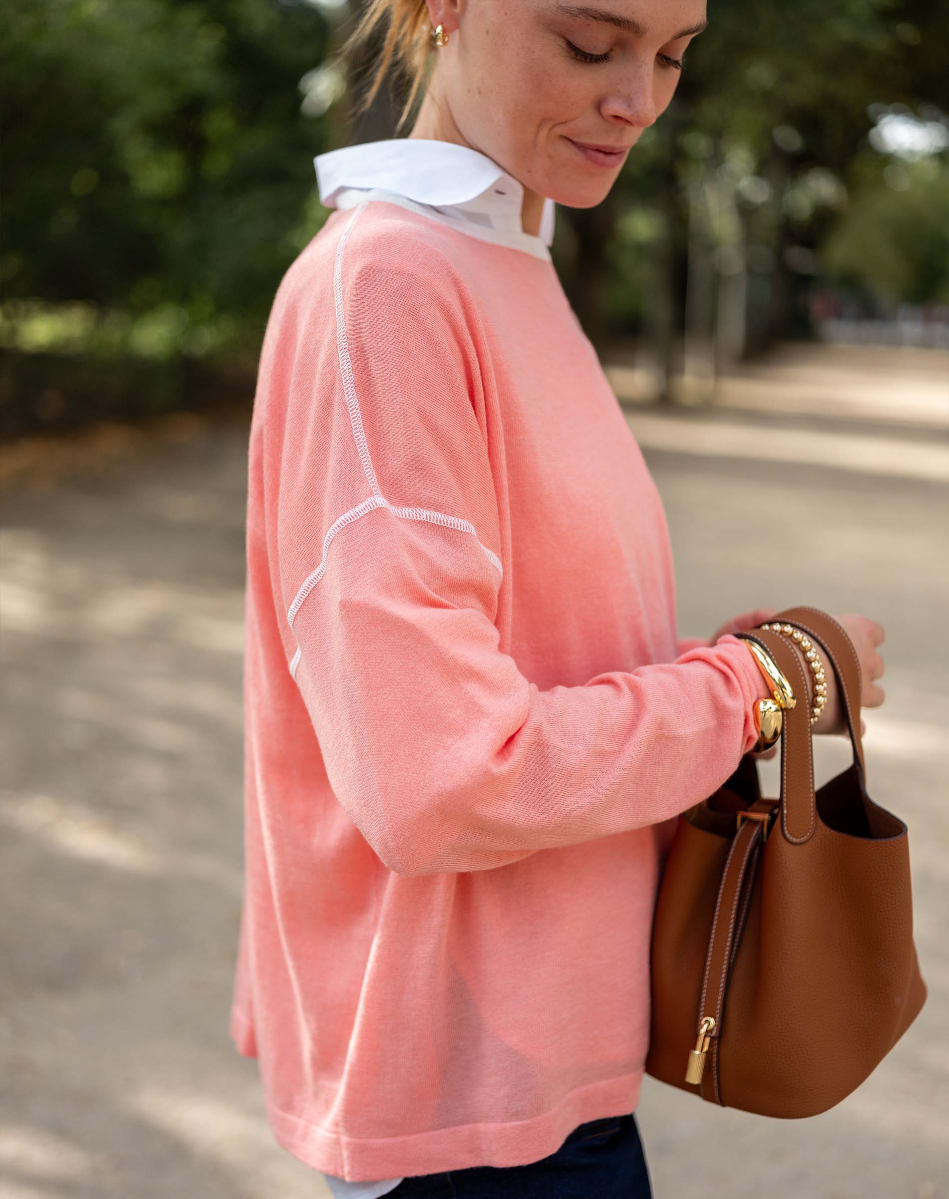 Woman wearing a pink sweater and holding a brown handbag outdoors.