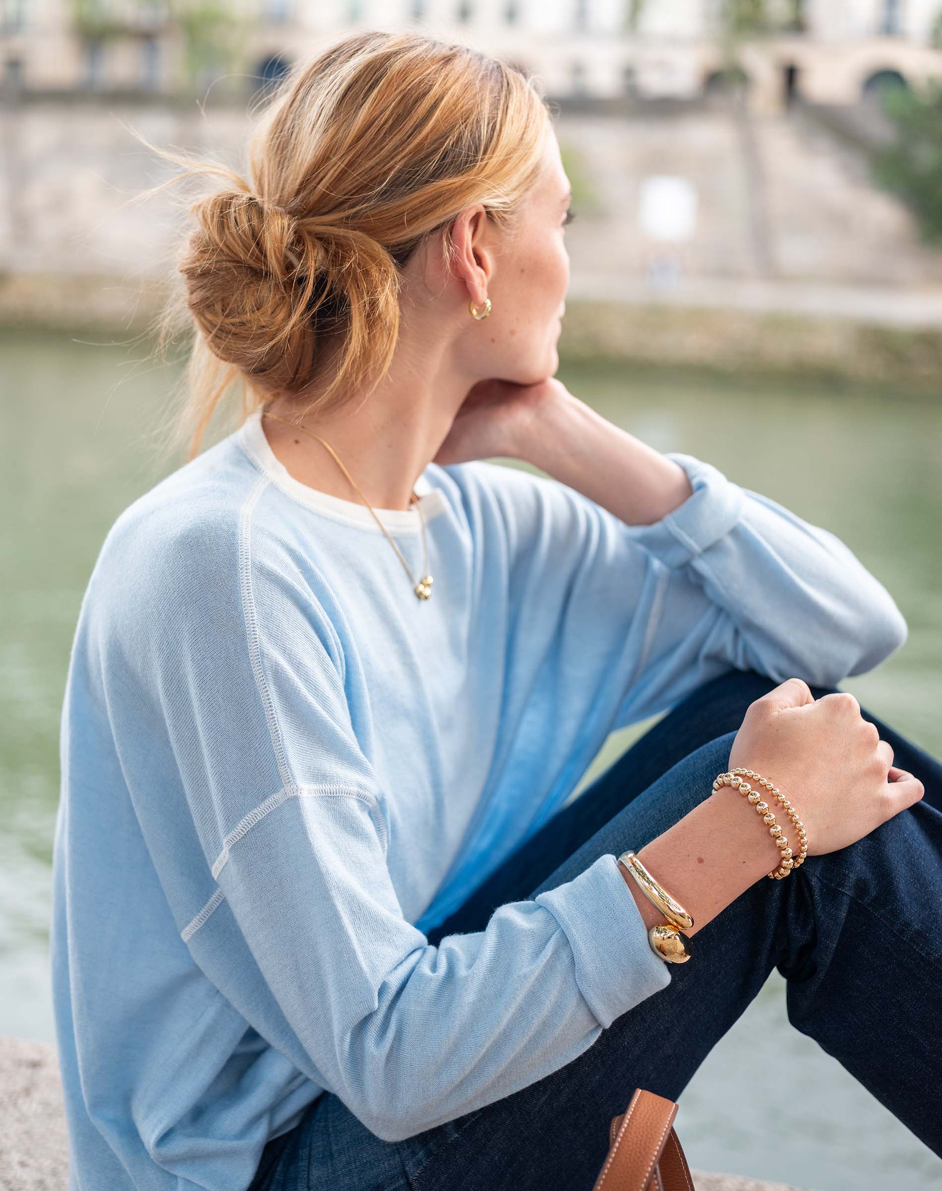 Woman sitting by a waterfront wearing a light blue shirt and dark jeans, with a blurred background.