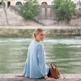 Woman in a light blue dress sitting by a river with a brown handbag.