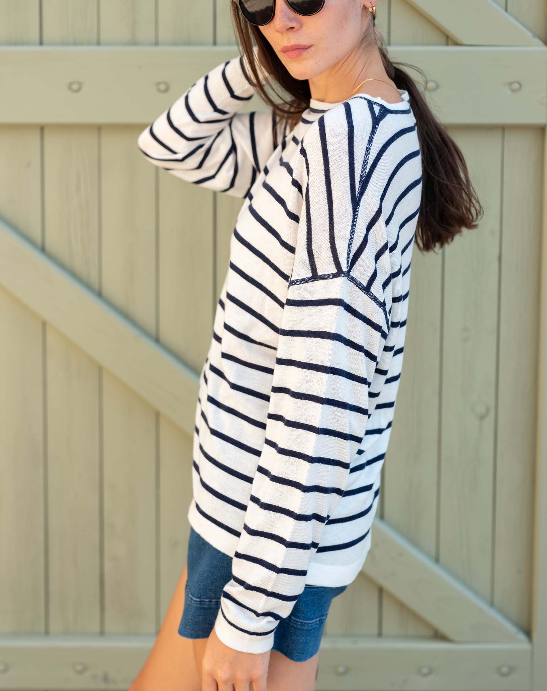 Woman wearing a striped long-sleeve shirt and denim shorts against a wooden fence.
