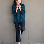 Woman in teal sweater and dark pants standing against a plain wall.