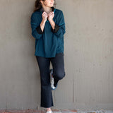 Woman in teal sweater and dark pants standing against a plain wall.