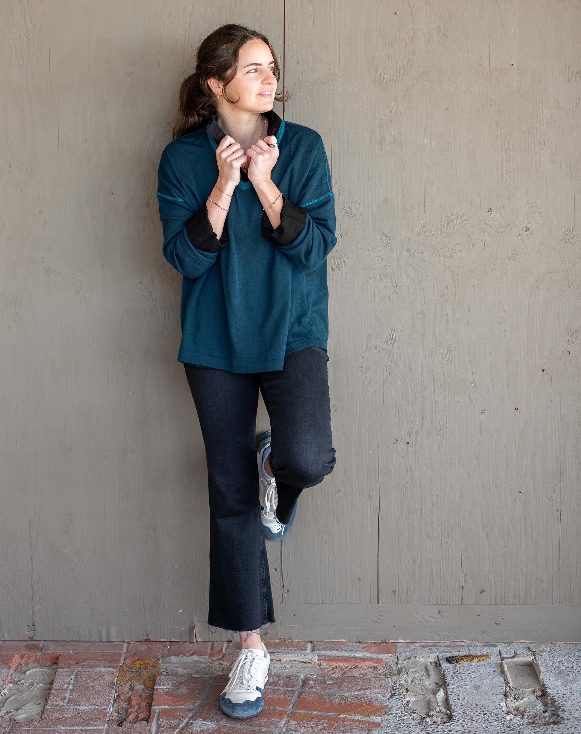 Woman in teal sweater and dark pants standing against a plain wall.