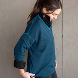 Woman wearing a teal sweater with black cuffs and collar against a gray wall.