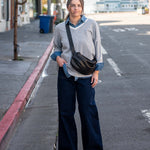 Woman standing on a street corner wearing a gray sweater and dark pants with a black bag.