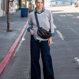 Woman standing on a street corner wearing a gray sweater and dark pants with a black bag.