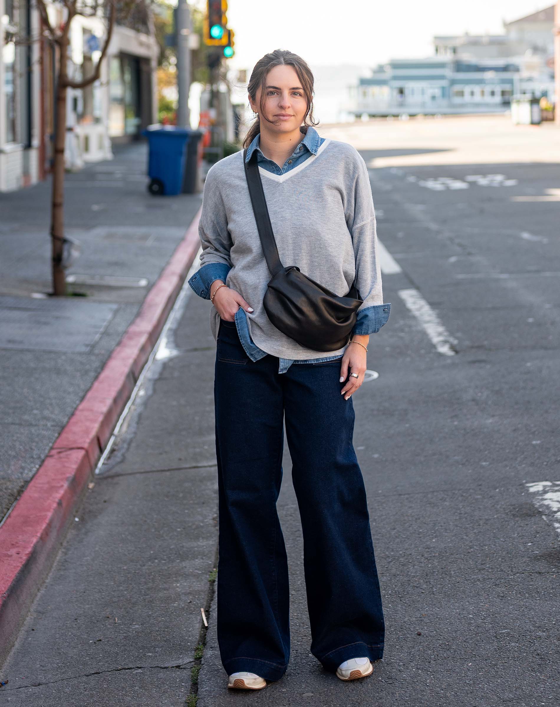 Woman standing on a street corner wearing a gray sweater and dark pants with a black bag.