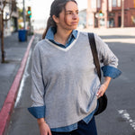 Woman standing on a street corner wearing a gray sweater with rolled-up sleeves and a denim shirt underneath.