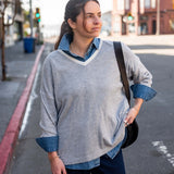 Woman standing on a street corner wearing a gray sweater with rolled-up sleeves and a denim shirt underneath.