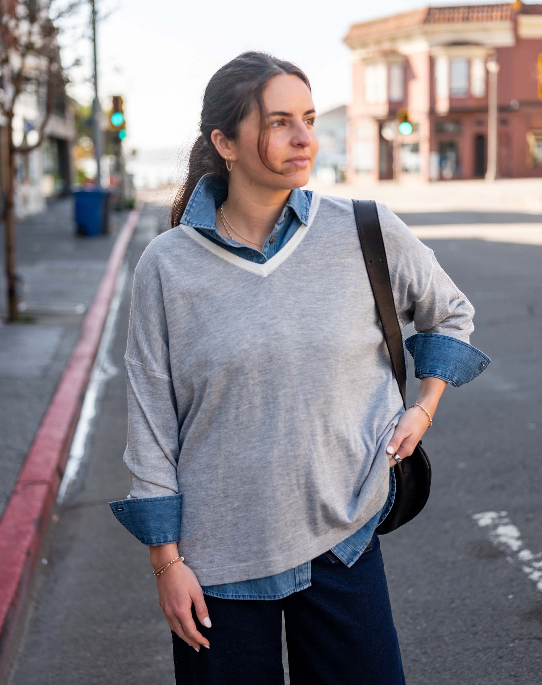 Woman standing on a street corner wearing a gray sweater with rolled-up sleeves and a denim shirt underneath.