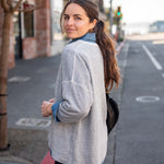 Woman standing on a street, wearing a light gray jacket and dark pants.