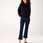 Woman showcasing Mersea moody blue Nomad cropped mini boot-cut jeans standing against white background