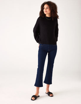 Woman showcasing Mersea moody blue Nomad cropped mini boot-cut jeans standing against white background