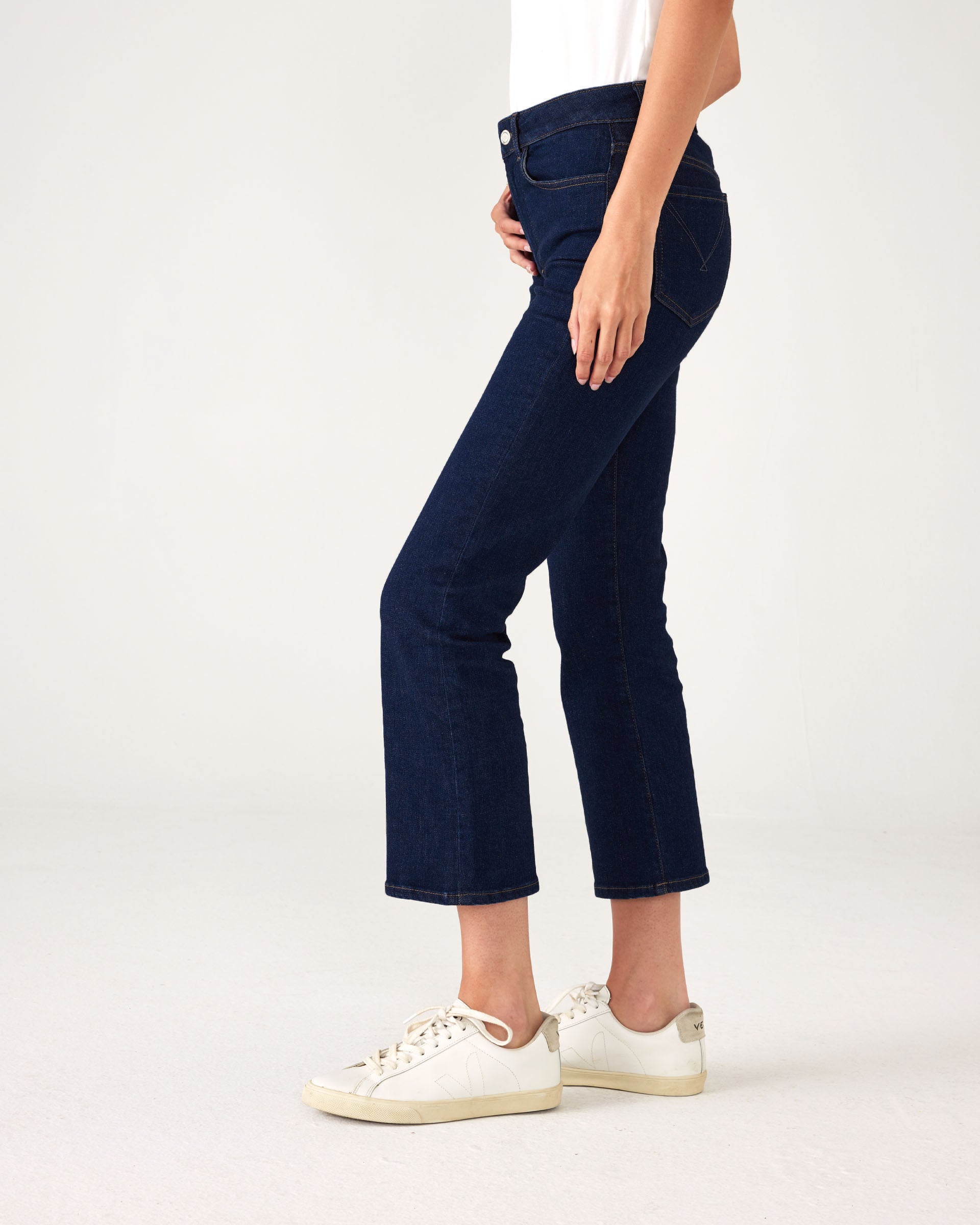 lower body side view of Woman showcasing Mersea moody blue Nomad cropped mini boot-cut jeans standing on white background