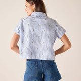 Woman wearing a blue and white striped shirt with floral patterns and denim shorts on a beige background