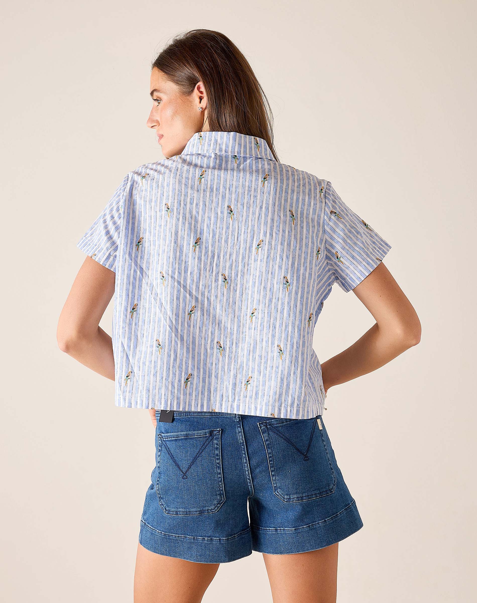 Woman wearing a blue and white striped shirt with floral patterns and denim shorts on a beige background