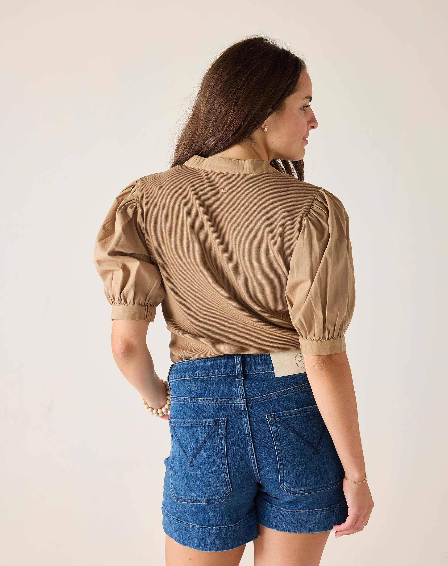 Woman wearing a beige blouse with puffed sleeves and blue denim shorts on a plain background