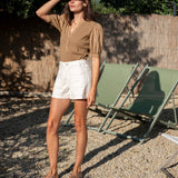 Woman in a brown shirt and white shorts standing outdoors with green chairs in the background.