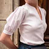 Woman wearing a white blouse with puffed sleeves and a black bag, standing against a stone wall.