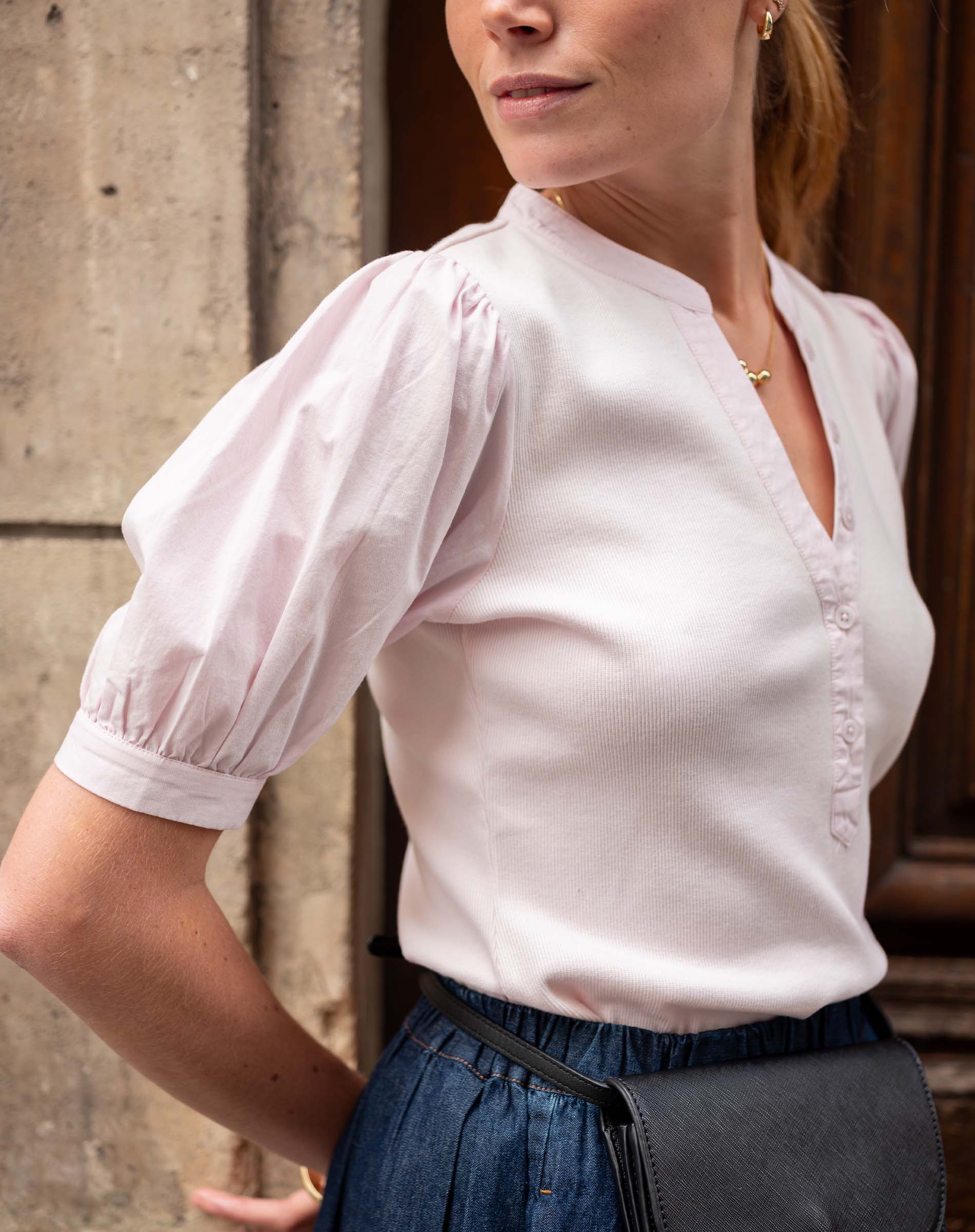 Woman wearing a white blouse with puffed sleeves and a black bag, standing against a stone wall.