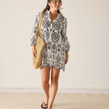 Woman wearing a patterned dress holding a straw bag against a plain background