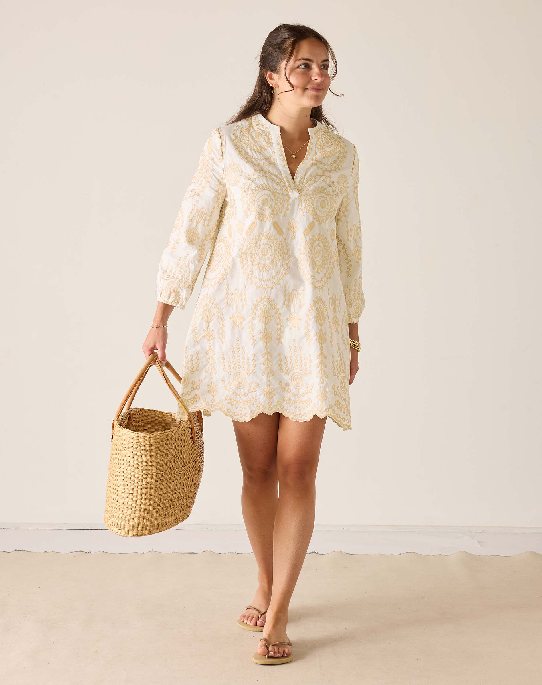 Woman wearing a light-colored dress holding a woven bag against a plain background