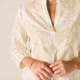 Close-up of a person wearing a white blouse with gold patterns on a plain background