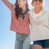 Two women standing on a beach, wearing striped shirts and sunglasses, with a clear blue sky in the background.
