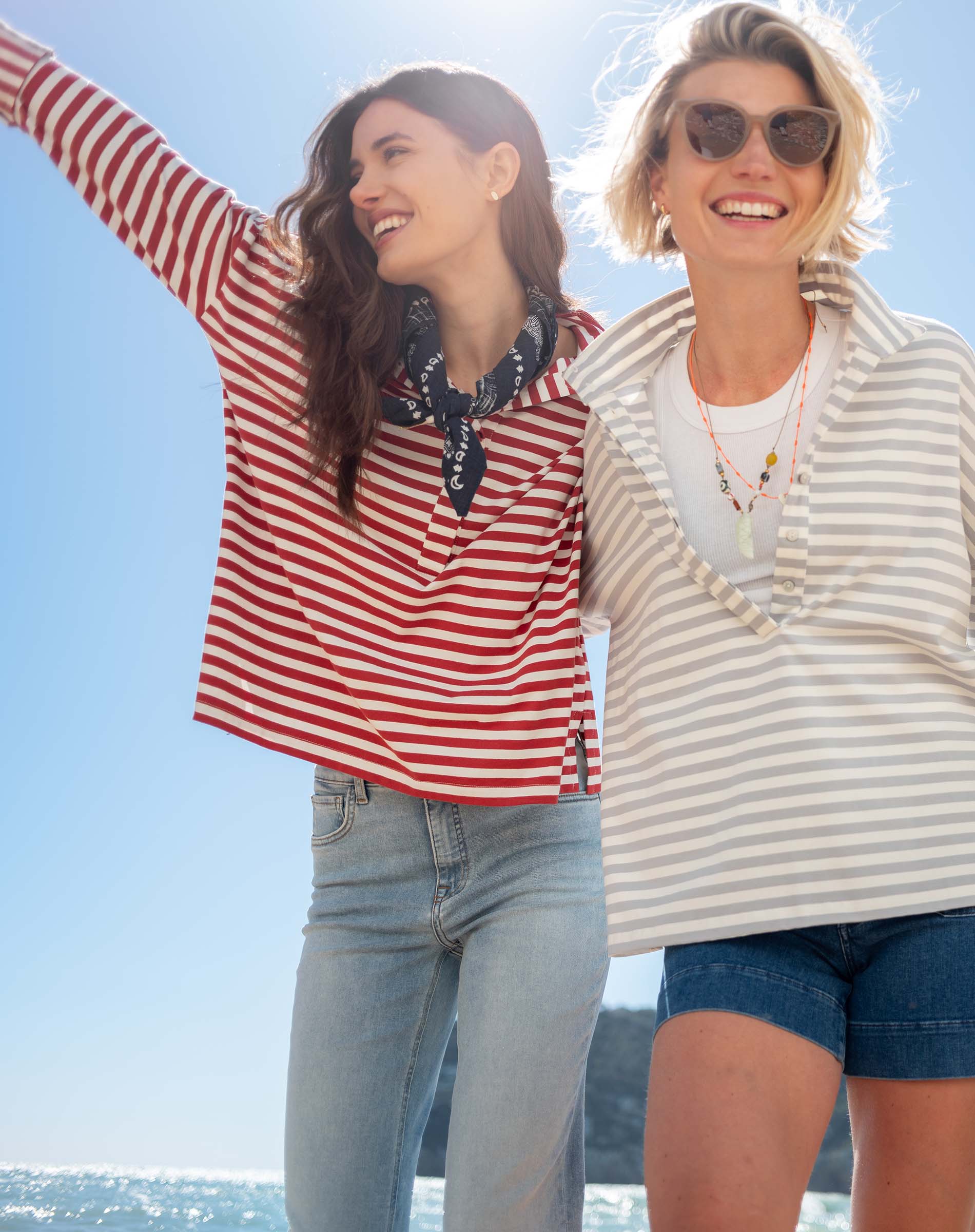Two women standing on a beach, wearing striped shirts and sunglasses, with a clear blue sky in the background.
