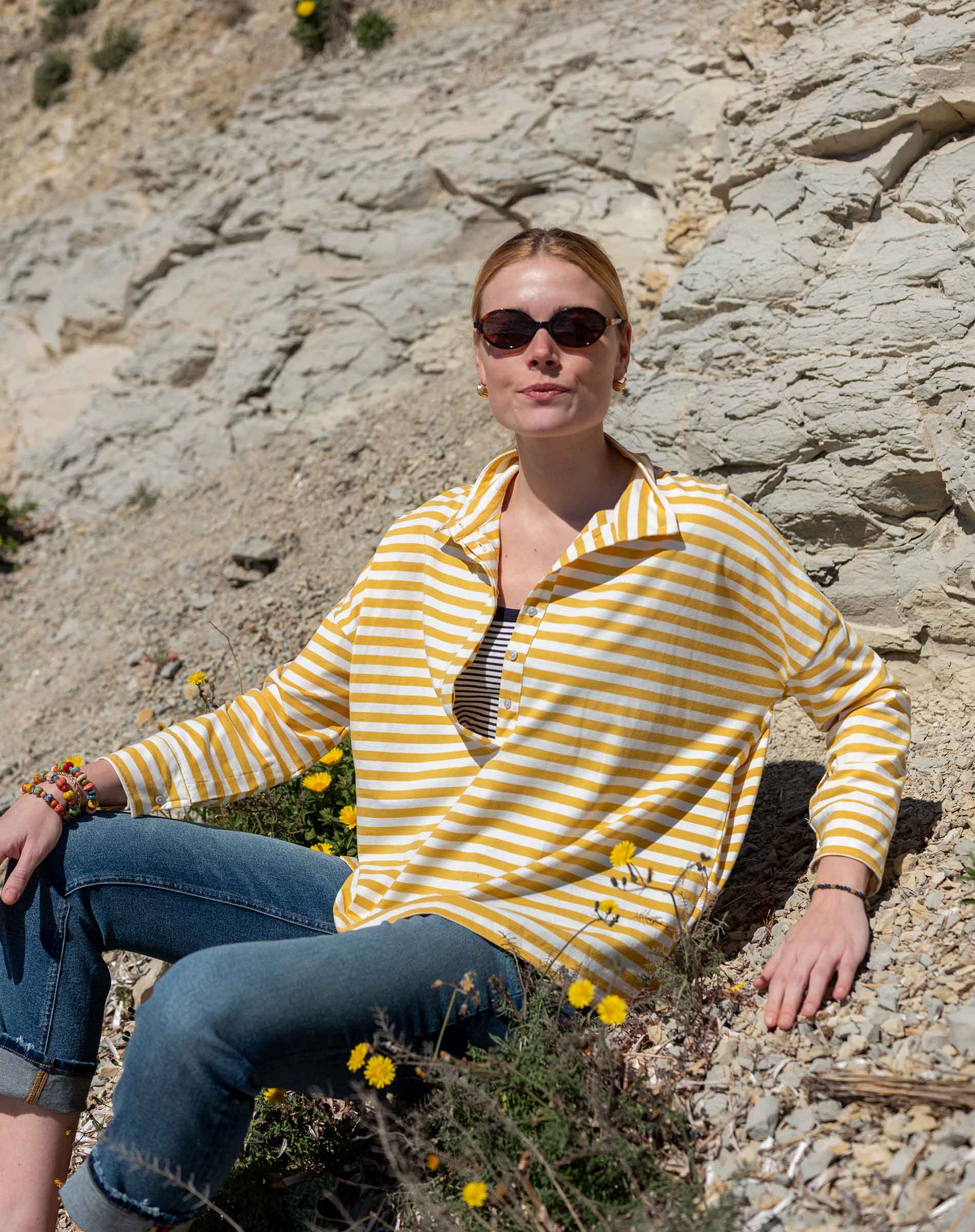 Woman in a yellow and white striped shirt sitting on rocky terrain with wildflowers.