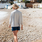 Woman in a striped shirt and shorts walking on a pebbly beach with buildings in the background.